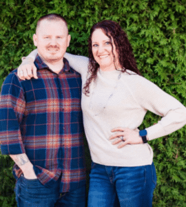 A smiling couple posing with arms around each other in front of a green hedge.